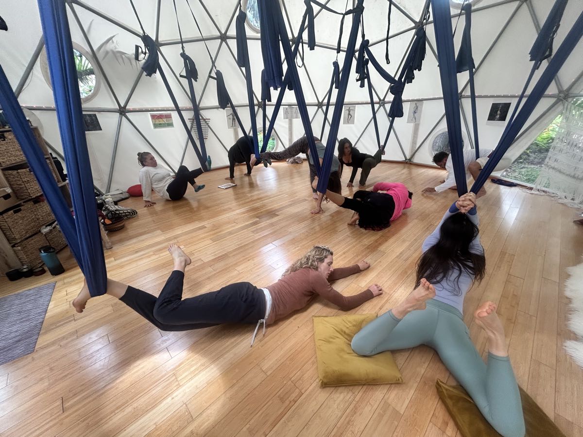 Several people together inside the Aum Dome, each one working with a hanging blue swing, some with a leg or both arms cradled in the fabric and others stretched out on the wood floor beside them.