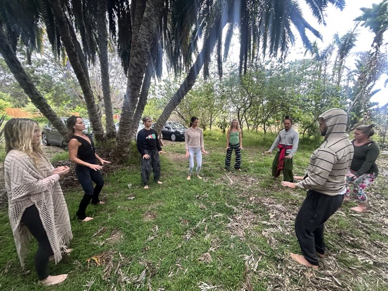 A small group standing barefoot on the grass in a wide circle under the palm trees outside the Aum Dome, arms lifted and open in a slow shared movement.