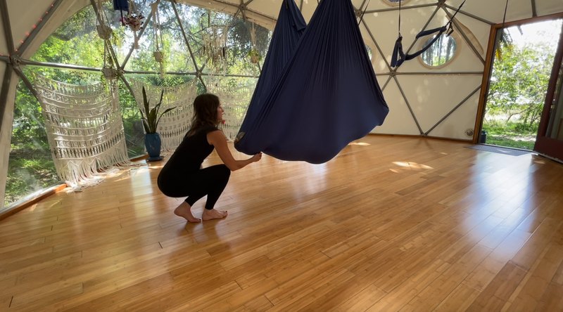 Maura Rassman crouched low beside a swing hanging heavy with a client held inside the fabric, her hands at the head end, sunlight falling across the bamboo floor of the Aum Dome